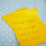 Une note manuscrite en coréen écrite sur des feuilles de papier jaune lignées posées sur une surface à pois bleus.