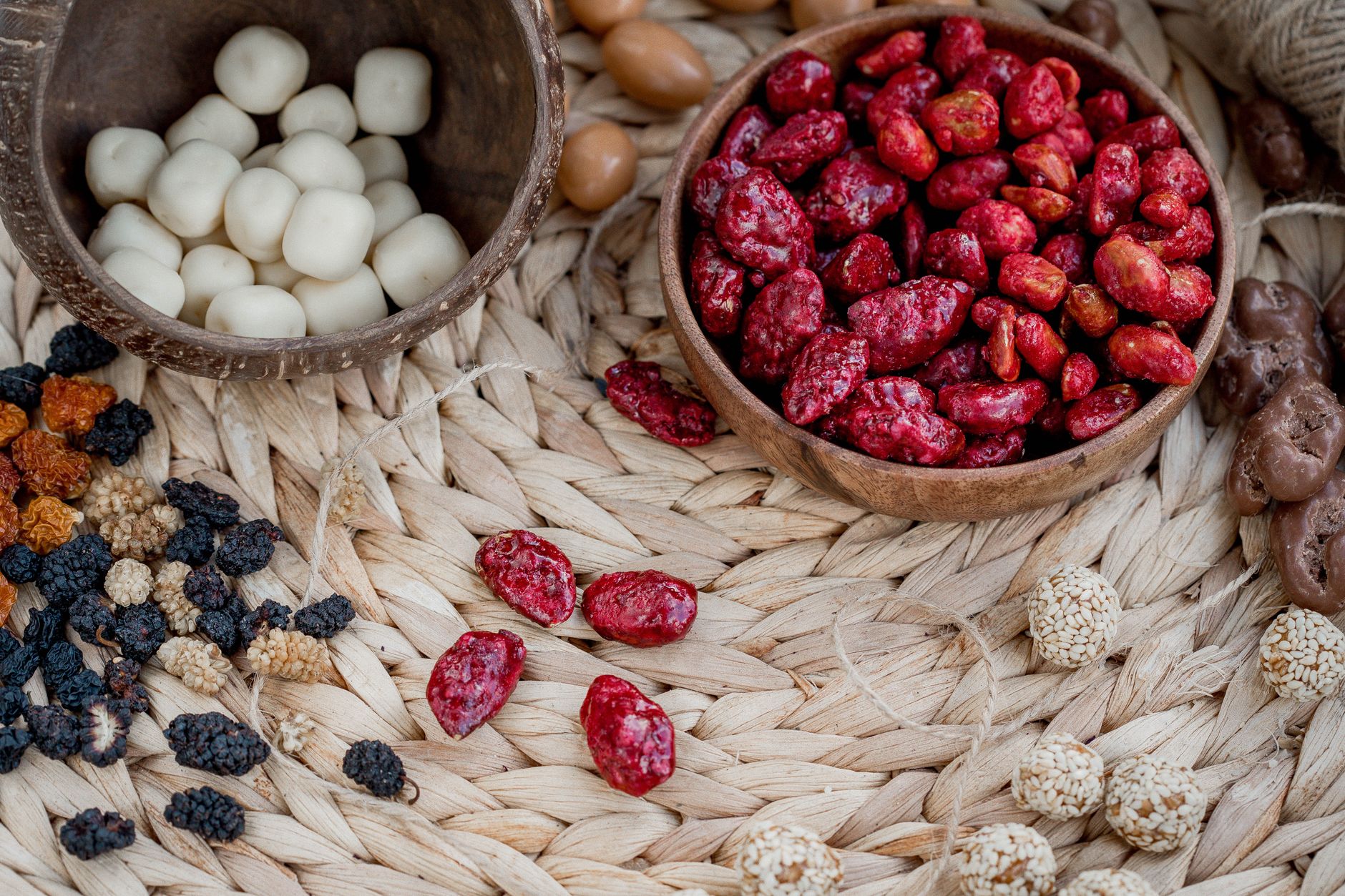 Des bols en bois remplis d'amandes dragéifiées rouges et blanches disposés sur un napperon en osier avec d'autres friandises.