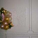 Un bouquet de ballons dorés et roses en forme d'étoiles et ronds devant un mur blanc mouluré.