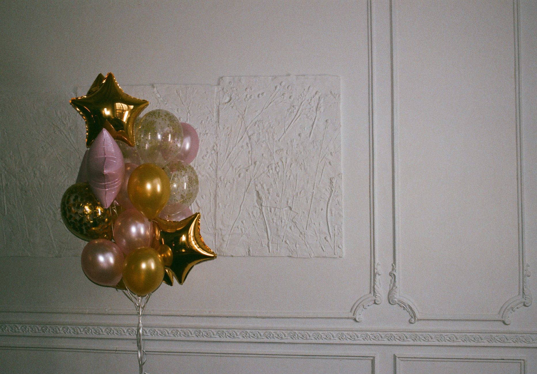 Un bouquet de ballons dorés et roses en forme d'étoiles et ronds devant un mur blanc mouluré.