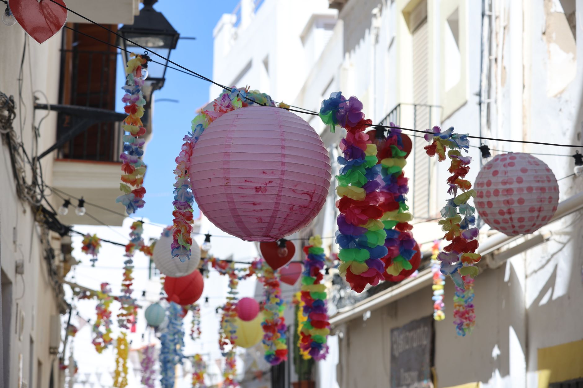 Décorations festives composées de lampions en papier et de guirlandes de fleurs colorées suspendues dans une rue étroite.