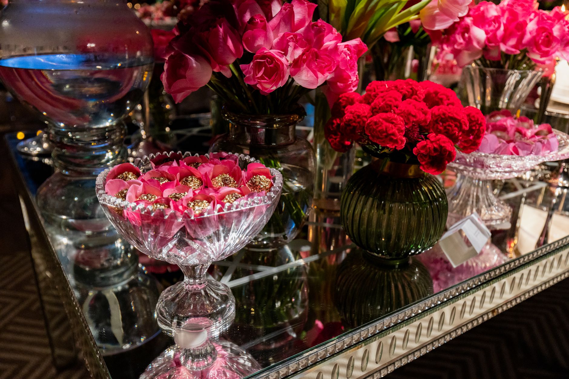 Une table de fête élégante ornée de vases en cristal, de fleurs roses et rouges, et de petites douceurs emballées dans du papier doré.