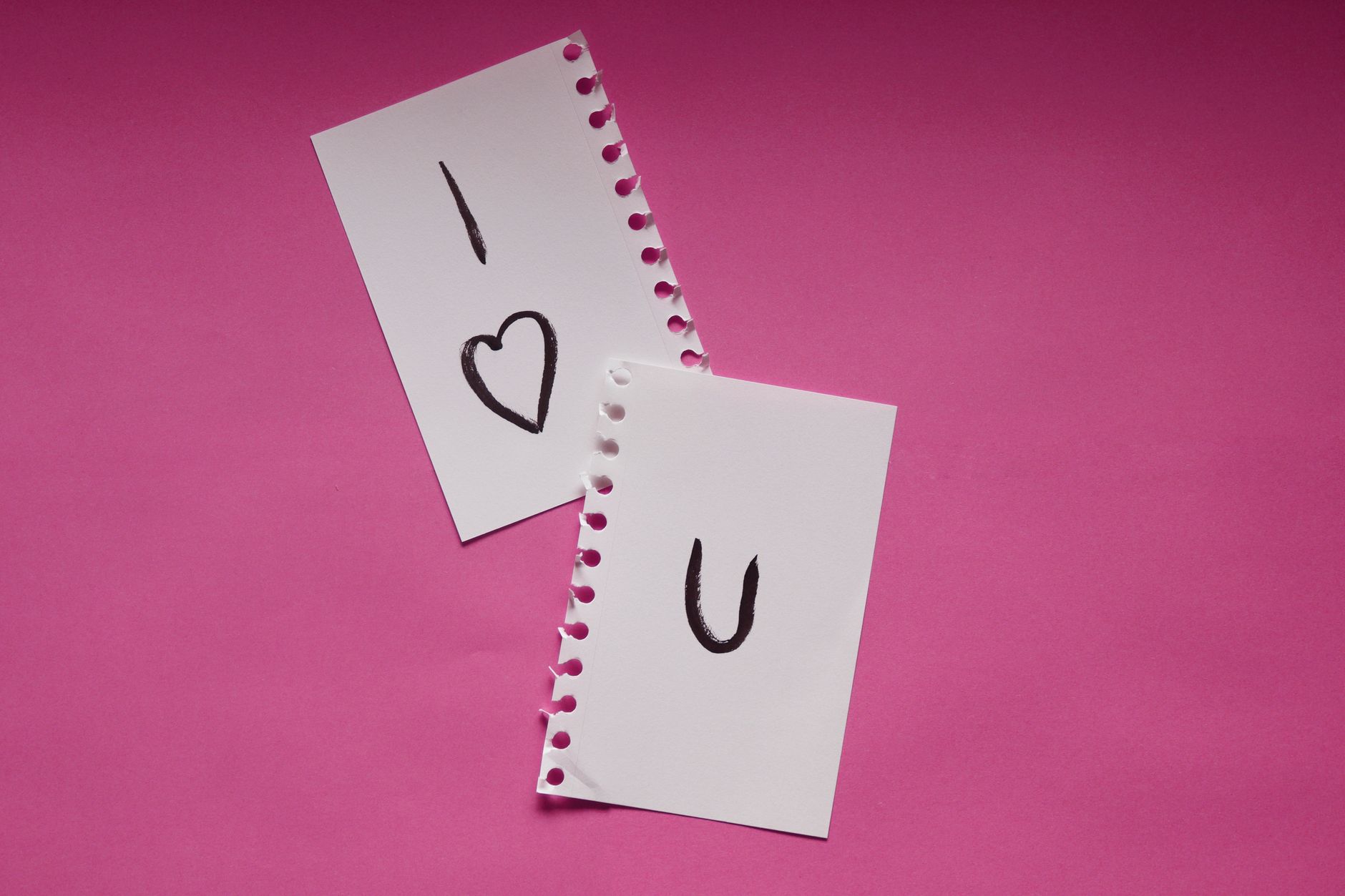 Deux morceaux de papier déchirés d'un carnet à spirales sur un fond rose, formant le message manuscrit I love you avec un cœur.